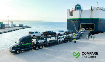 Editorial photograph of a car carrier truck loaded with American vehicles parked next to a cargo ship at a port during sunrise.