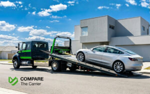 Professional flatbed tow truck winching a silver electric sedan onto the deck on a bright, clean residential street.
